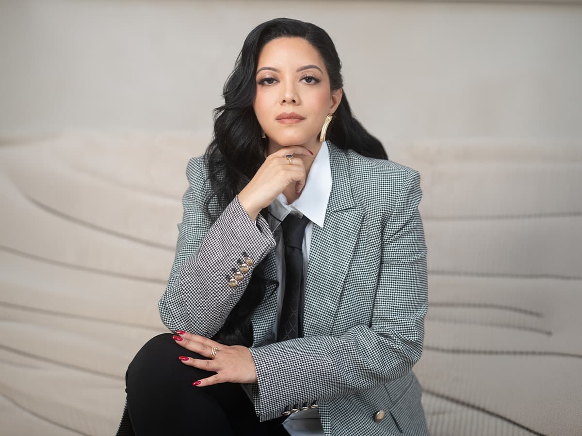 Priscilla Laclé seated on steps in a checked blazer, resting her chin on her hand